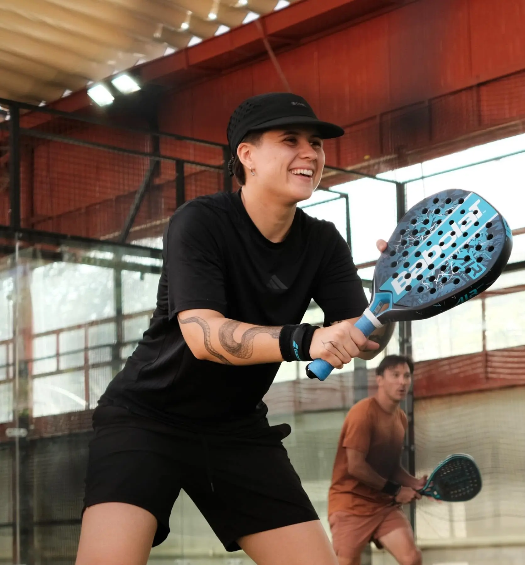 Jenny mid-rally on an indoor padel court, paddle raised, smiling, opponent visible in the background.